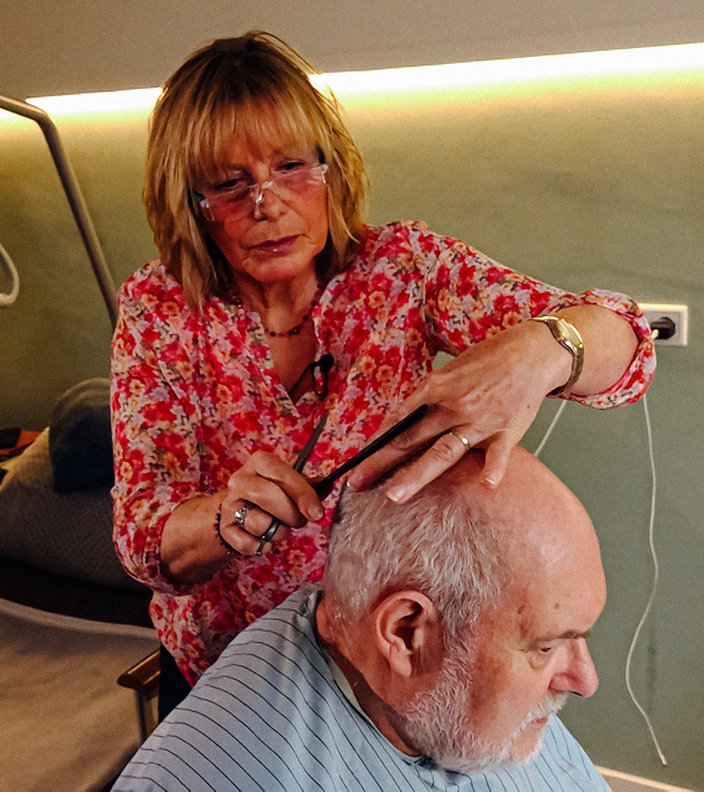 Im Vordergrund sitzt Hospizpatient Jürgen mit einer Friseurschürze auf einem Rollstuhl. Dahinter steht Marion Hebel und schneidet ihm die Haare. Im Hintergrund ist das Hospizbett zu sehen.