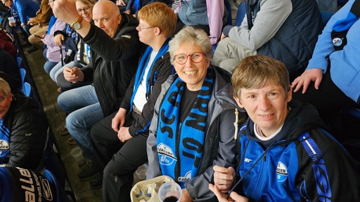 Auf dem Bild sind mehrere Menschen in einem Stadion zu sehen. Sie sitzen blauen Sitzschalen aus Plastik.