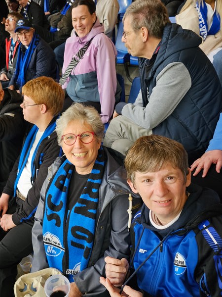 Auf dem Bild sind mehrere Menschen in einem Stadion zu sehen. Sie sitzen blauen Sitzschalen aus Plastik.