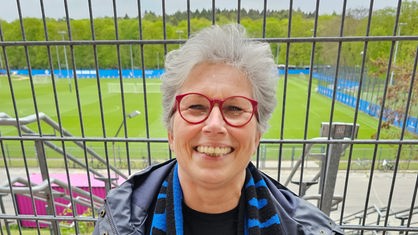 Auf dem Bild stehe eine Frau mt roter Brille und weißen Haaren. Sie trägt einen blauen Schal des SC Paderborn. Im Hintergrund ist ein Fußballfeld zu sehen.