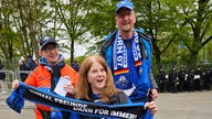 Auf dem Bild sind drei Menschen zu sehen. Sie sind Fans des SC Paderborn und tragen blaue Triktots und Schals. Eine Person sitzt im Rollstuhl.
