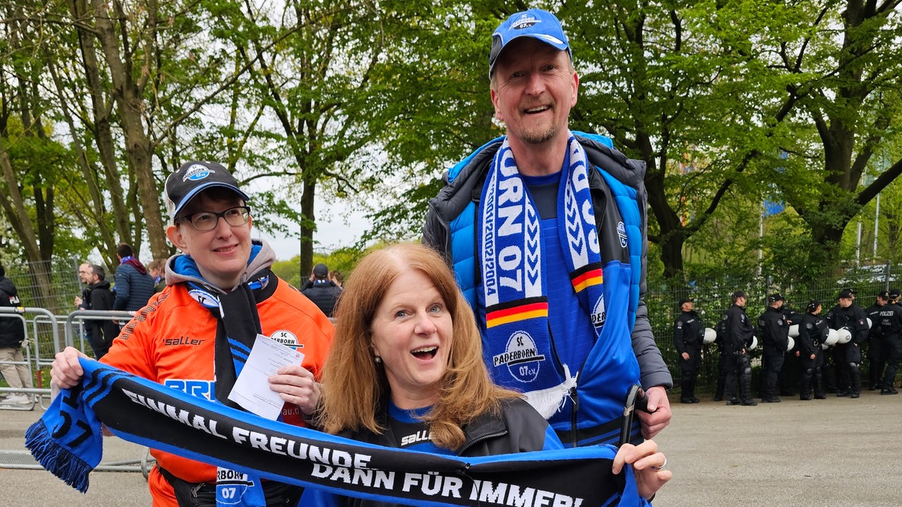Auf dem Bild sind drei Menschen zu sehen. Sie sind Fans des SC Paderborn und tragen blaue Triktots und Schals. Eine Person sitzt im Rollstuhl.