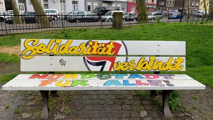 Auf einer weißen Bank steht in bunter Schrift: Solidarität verbindet, Nordstadt für alle.