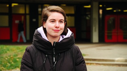 Eine junge Frau in schwarzer Winterjacke und braunen glatten Haaren vor einer Schule