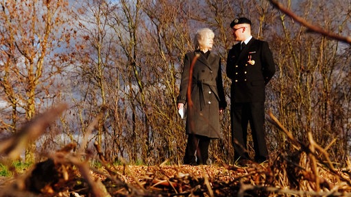 Ein Mann und eine Frau stehen nebeneinander an einem spätherbstlichen Wald und schauen sich an. 