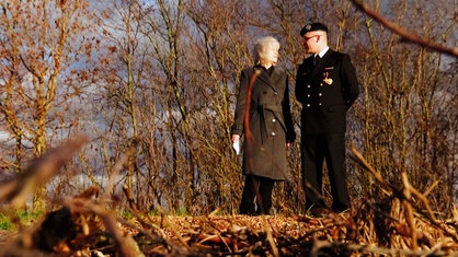 Ein Mann und eine Frau stehen nebeneinander an einem spätherbstlichen Wald und schauen sich an. 