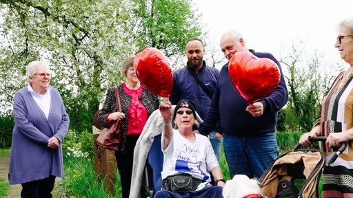 Martina sitzt im Rollstuhl. Um sie herum stehen Familie und Freunde mit Herzballons zum Geburtstag.