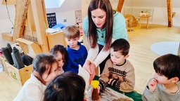 Kita-Kinder sitzen um einen Tisch. Auf dem Tisch ist ein Experiment mit einem kleinen Plastikvulkan aufgebaut. Nina Beyer ist über dem Tisch gebeugt und leitet die Kinder an.