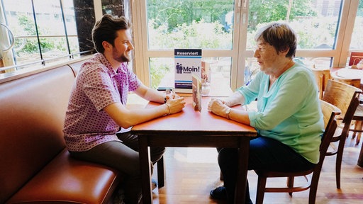 Ein Student und eine Seniorin sitzen sich in einem Café gegenüber und unterhalten sich.