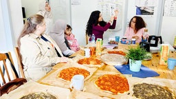 Bilge Akbulut hält einen Zettel hoch auf dem verschiedene Vokabeln erklärt werden. Die Teilnehmerin hören ihr interessiert zu. Auf dem Tsich stehen mehrere libanesische Pizzen.