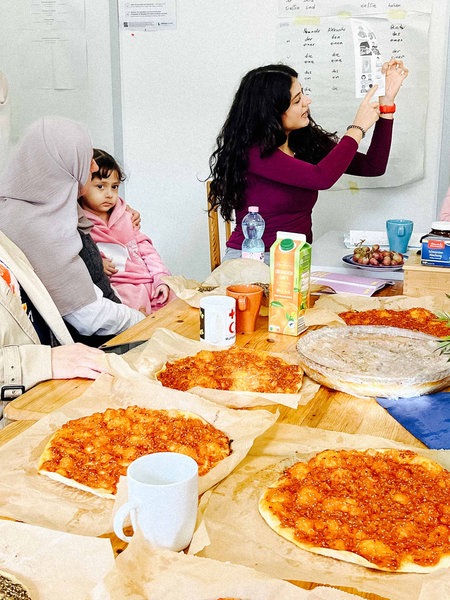 Bilge Akbulut hält einen Zettel hoch auf dem verschiedene Vokabeln erklärt werden. Die Teilnehmerin hören ihr interessiert zu. Auf dem Tsich stehen mehrere libanesische Pizzen.