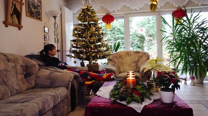 Sarah Easter sitzt im Wohnzimmer auf dem Sofa unter ihrer Decke. Neben ihr der Weihnachtsbaum.