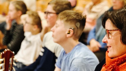 Mehrere gehörlose Kinder sitzen nebeneinander. Ein Kind trägt ein Hörgerät. Ganz rechts sitzt eine Mutter.