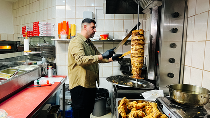 Ein Mann mit dunklen Haaren schneidet mit einem großen Messer Fleisch von einem Schawarma-Spieß