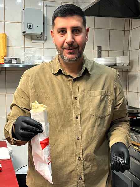 Ein Mann mit dunklen Haaren und Bart steht in einer Restaurantküche und hält ein Schawarma in seiner Hand 