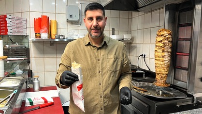 Ein Mann mit dunklen Haaren und Bart steht in einer Restaurantküche und hält ein Schawarma in seiner Hand 