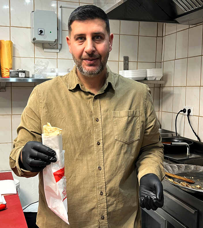 Ein Mann mit dunklen Haaren und Bart steht in einer Restaurantküche und hält ein Schawarma in seiner Hand 