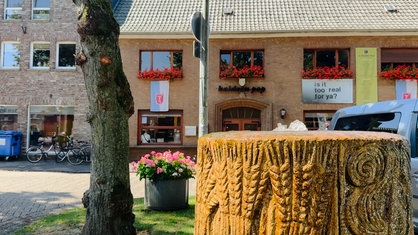 Im Vordergrund ist ein Baumstamm zu sehen, im Hintergrund erkennt man die Außenfassade eines Cafés.