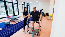 Ein Mann übt mit einer Art Rollator, zu gehen. Eine Physiotherapeutin steht neben ihm.