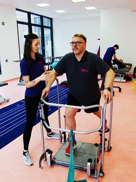 Ein Mann übt mit einer Art Rollator, zu gehen. Eine Physiotherapeutin steht neben ihm.