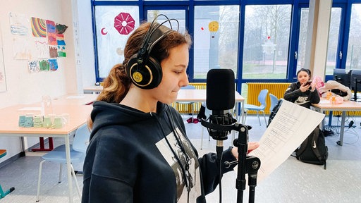 Cheyenne Erler steht beim Rap-Workshop im Klassenzimmer mit Kopfhörern vor einem Mikrofon. In der Hand hat sie einen Zettel mit ihrem Text.