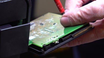 Eine Hand hält einen grüne Computer-Platine und arretiert die Spitze eines Schraubendrehers auf ihr