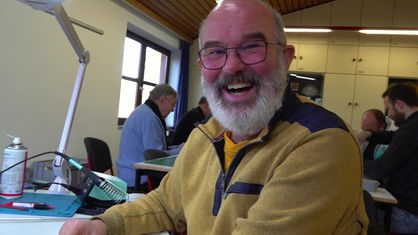 Ein Mann mit grauem Bart, schwarzer Brille, Glatze und gelbem Pullover lacht in die Kamera