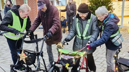 Zwei Personen wollen mit einem Rollator eine Treppenstufe hoch. Ihnen helfen zwei weitere Personen 