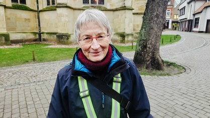 Eine Frau mit kurzen grauen Haaren steht vor einer Kirche und lächelt 