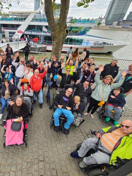 Eine Gruppe von Menschen am Rheinufer, im Hintergrund liegt ein Schiff. Einige der Menschen sitzen in Rollstühlen.