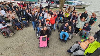 Eine Gruppe von Menschen am Rheinufer, im Hintergrund liegt ein Schiff. Einige der Menschen sitzen in Rollstühlen.