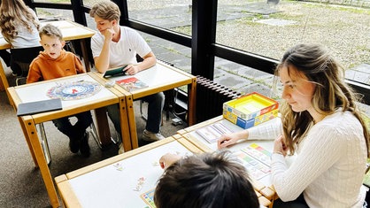 Zwei Schüler und eine Schülerin sitzen an Tischen in einer Schulbibliothek und lernen.