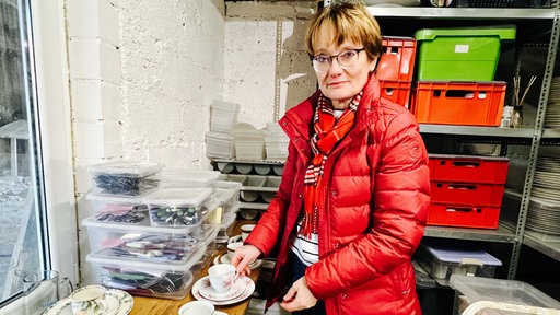 Simone Böhm steht in ihrem Geschirrlager vor einem Tisch auf dem verschiedene Kaffeetassen aufgereicht sind. Um sie herum stehen weitere Boxen mit Messern, Gabeln und Tellern.