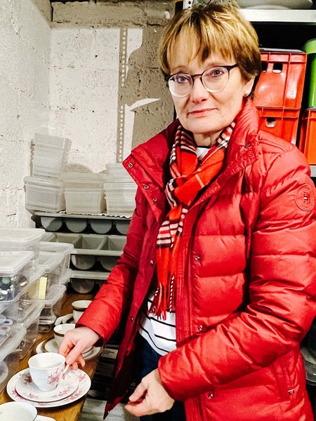 Simone Böhm steht in ihrem Geschirrlager vor einem Tisch auf dem verschiedene Kaffeetassen aufgereicht sind. Um sie herum stehen weitere Boxen mit Messern, Gabeln und Tellern.