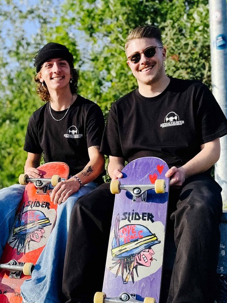 Fabio Navaro und Michael Franke sitzen auf einer Skateboardrampe, in der Hand halten sie ihre Skateboards