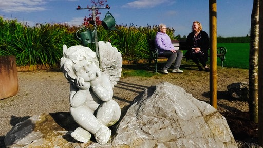 Zwei Frauen sitzen auf einem Friedhof auf einer Bank, im Vordergrund eine Engelsfigur