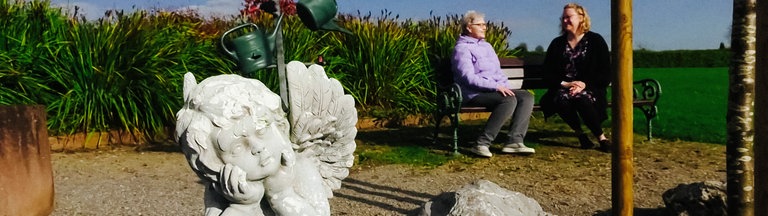 Zwei Frauen sitzen auf einem Friedhof auf einer Bank, im Vordergrund eine Engelsfigur