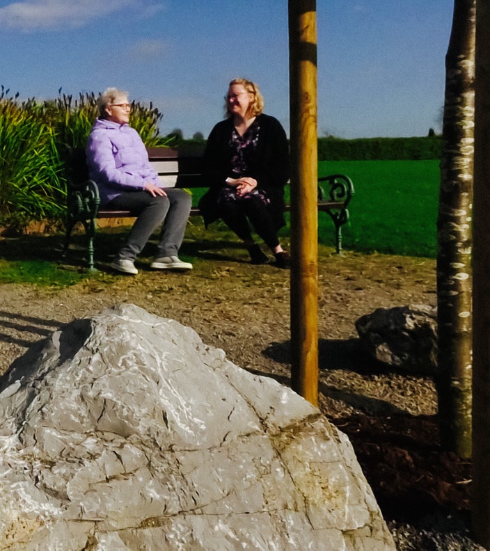 Zwei Frauen sitzen auf einem Friedhof auf einer Bank, im Vordergrund eine Engelsfigur