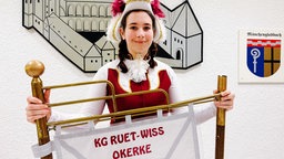 Vanessa Buch steht vor einer Wand an der eine Zeichnung einer Burg und das Wappen von Mönchengladbach hängt. In den Händen hält sie einen Banner mit der Aufschrift "KG Ruet-Wiss-Okerke". 