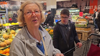 Monika Busch im Lebensmittelladen mit einem Einkaufswagen. Im Hintergrund steht ihr Helfer Fabian Reimann.