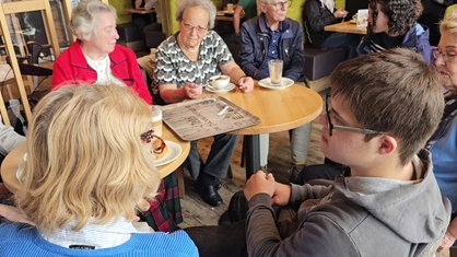 Ein junger Mann und mehrere ältere Frauen sitzen an einem Tisch in einem Café und sprechen miteinander.