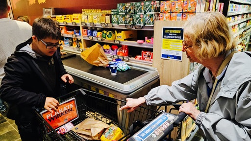 Ein Mann und eine Frau stehen in einem Supermarkt mit einem Einkaufswagen an einer Kasse.