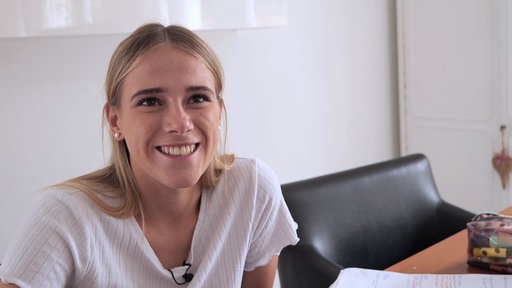 Eine junge Frau sitzt an einem Tisch. Sie ist blond und trägt ein weißes T-Shirt. Ihr Blick ist Richtung Kamera gerichtet.