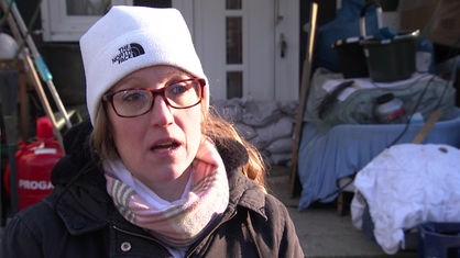 Eine Frau mit weißer Mütze und Brille steht vor einem Haus mit gestapelten Sandsäcken. 