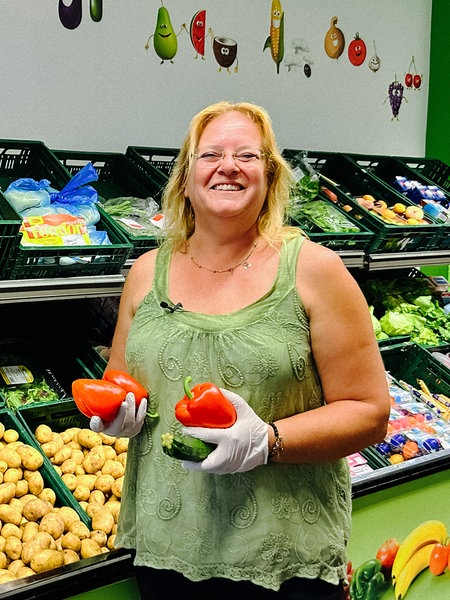 Eine Frau hält Gemüse in den Händen und posiert vor Kisten voller Gemüse und Kartoffeln.