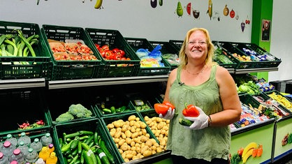 Eine Frau hält Gemüse in den Händen und posiert vor Kisten voller Gemüse und Kartoffeln.