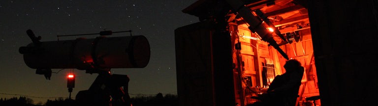 Hubertus Rieger schaut bei Nacht durch sein großes Teleskop. Dabei sitzt er zurückgelehnt in einem Stuhl. Im Hintergrund leuchtet der Innenraum der Mini-Sternwarte in einem orangen Licht.