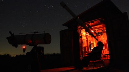 Hubertus Rieger schaut bei Nacht durch sein großes Teleskop. Dabei sitzt er zurückgelehnt in einem Stuhl. Im Hintergrund leuchtet der Innenraum der Mini-Sternwarte in einem orangen Licht.