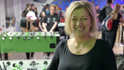 Eine Frau mit blonden, mittellangen Haaren lächelt in die Kamera. Im Hintergrund spielen Personen Tischfußball. 