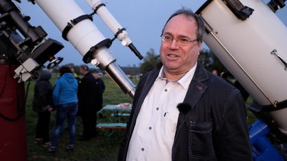 Hobbyastronom Hubertus Rieger steht lächelnd vor seinem Teleskop. Er trägt ein weißes Hemd und eine dunkle Jeans-Jacke.